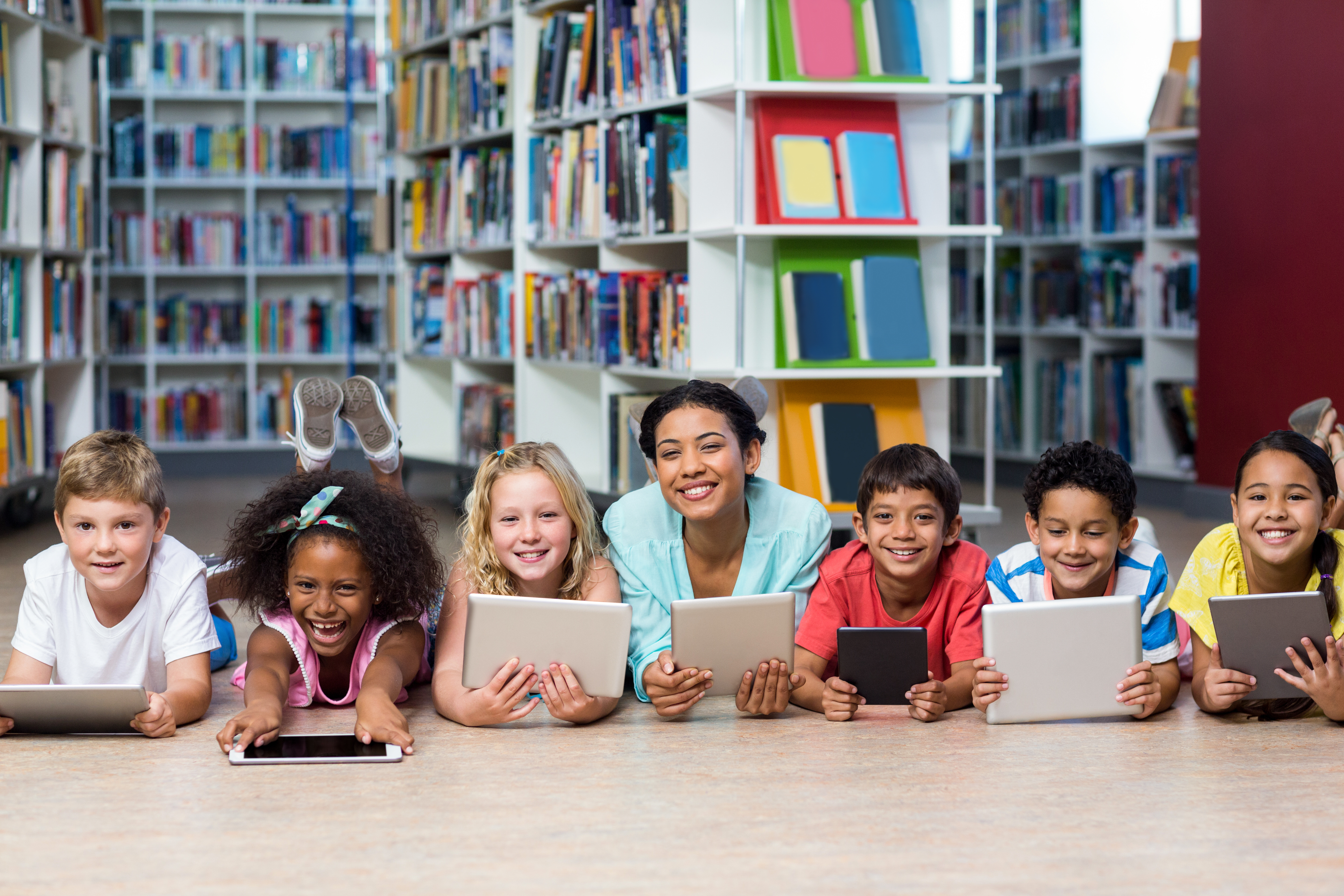 Elementary school teacher and her students lying on the floor of a library holding tablet devices