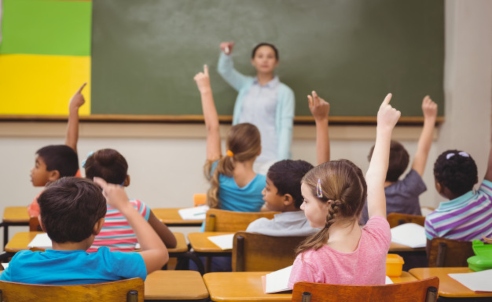 Children raising their hands in a classroom as the teacher selects a participant