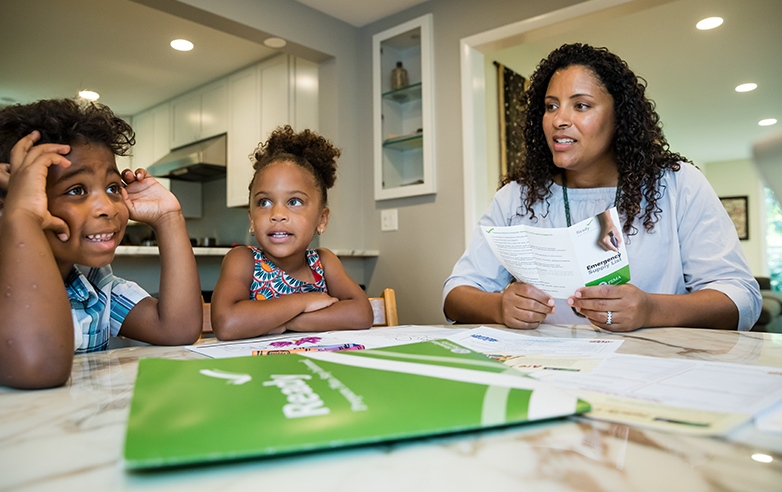 A woman is speaking with her two children at a table where Ready materials are spread out and being reviewed