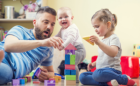A man in a striped shirt is playing with two children and creating a tower using wooden blocks