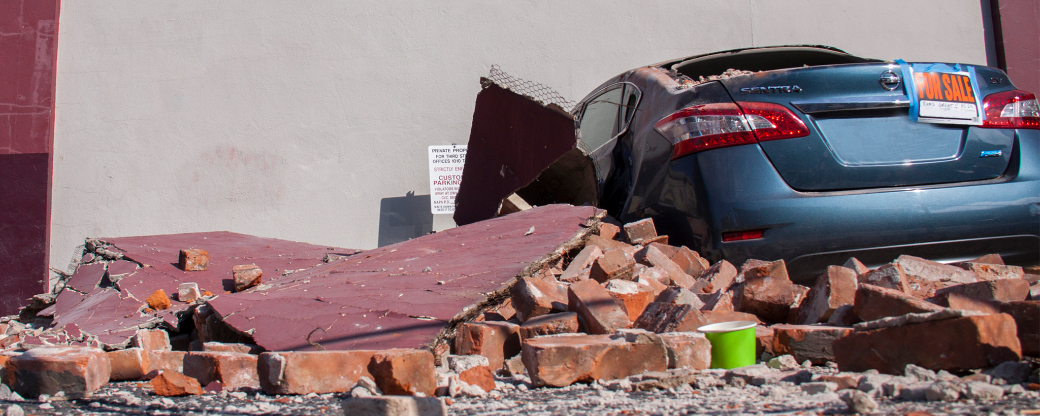 A car with a for sale sign heavily damaged by falling bricks and other debris during an earthquake