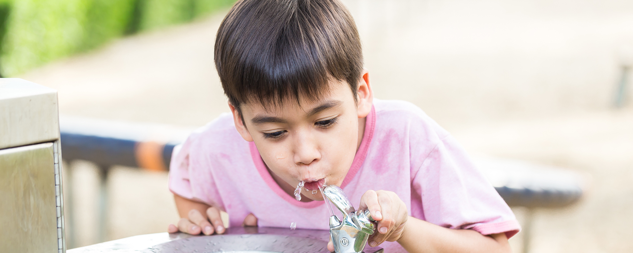 A boy in a pink shirt drinking from a water fountain outside