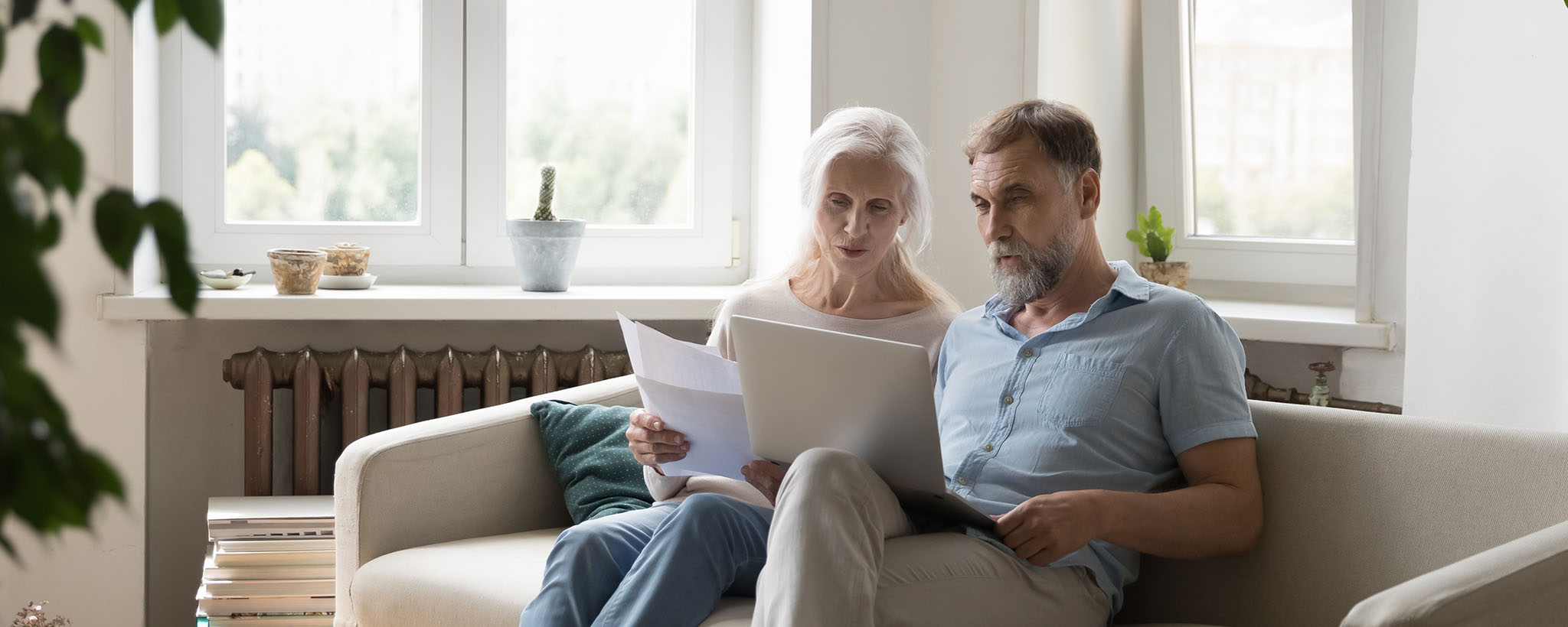 An older couple sit on the couch looking at their laptop and papers.