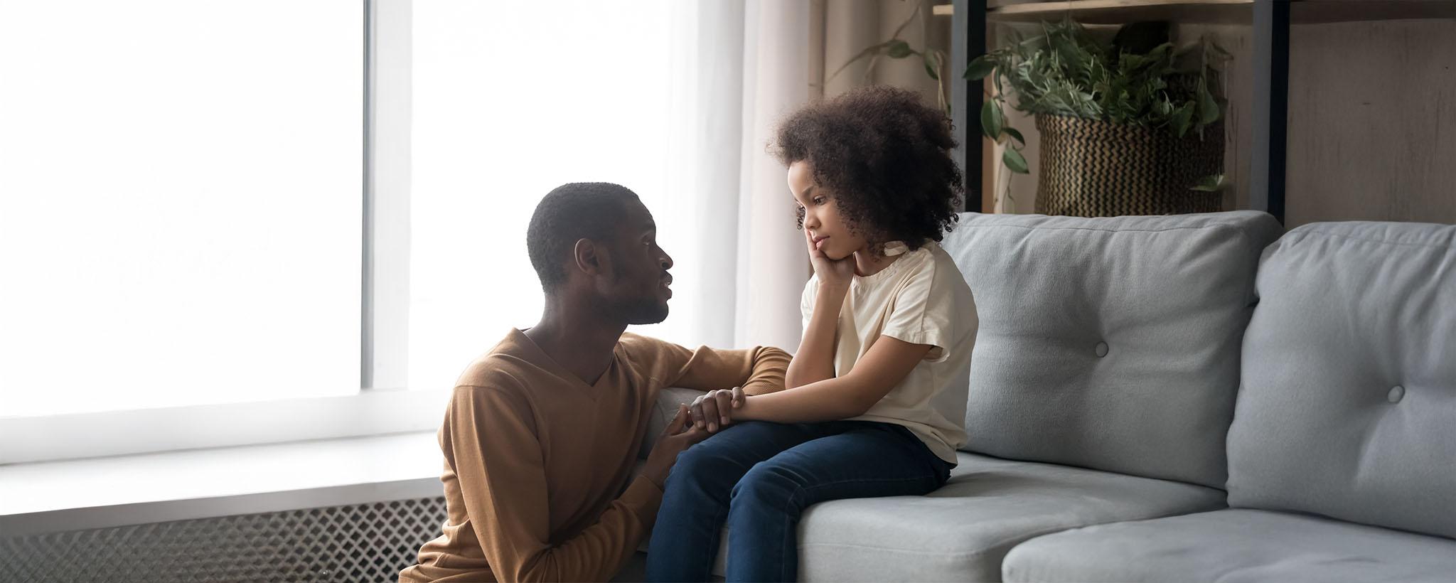 A father comforts his young upset daughter, sitting on the couch. 