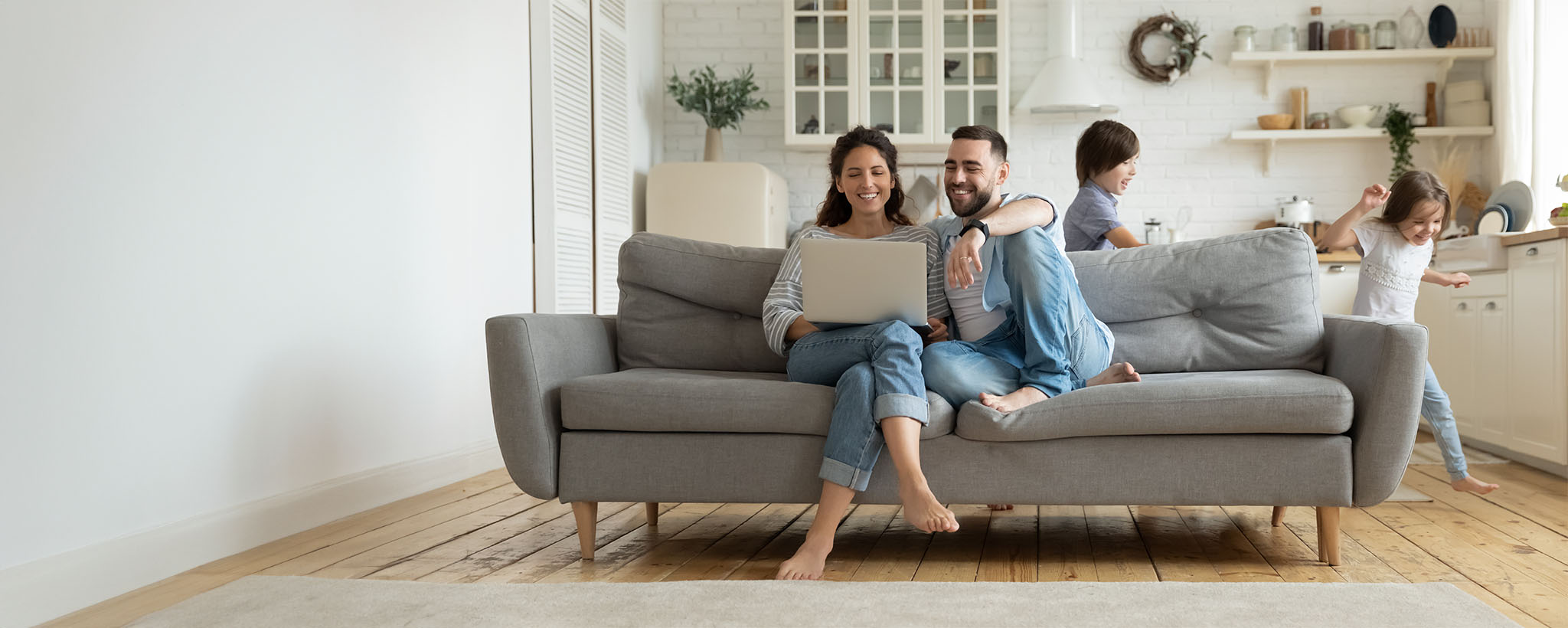 A mother and father sitting on the couch looking at a laptop while a school-aged girl and boy play around them. 