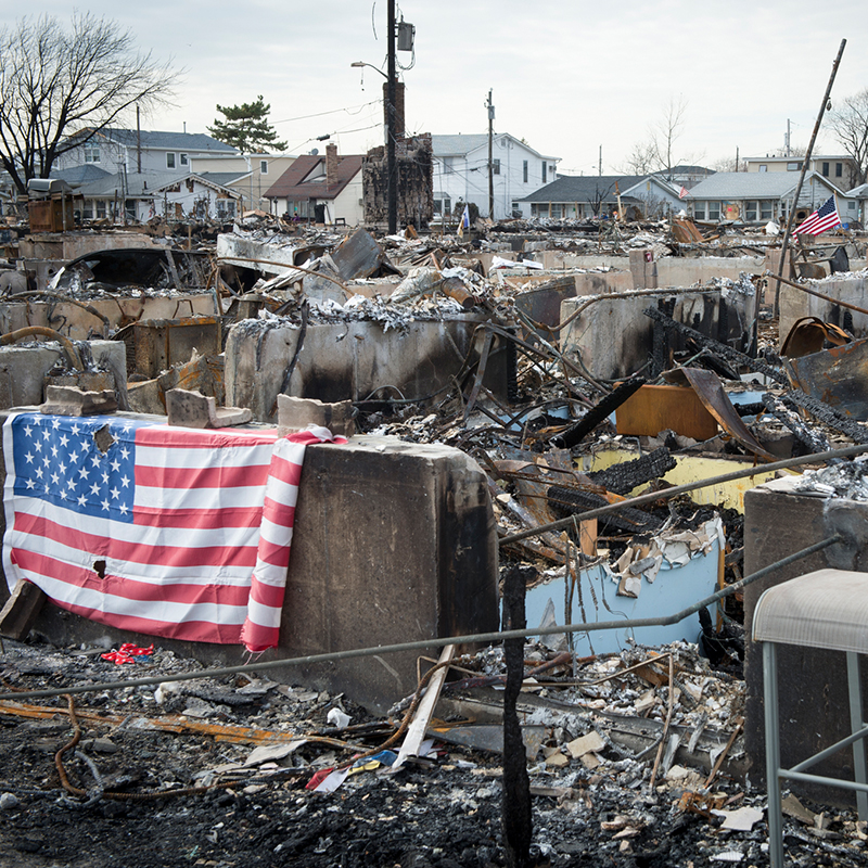 Decimated houses and rubble after hurricane Sandy, with an American flag drapped over the damage.