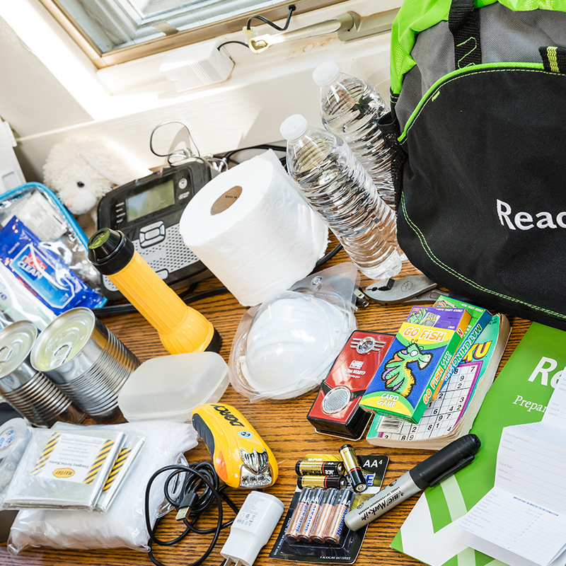 The items in an emergency supply kit spread out on a table including water bottles, toilet paper, and batteries