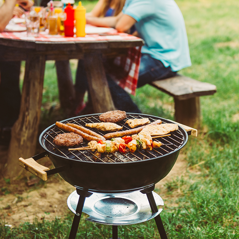 Una parrilla de barbacoa con hamburguesas y salchichas, la gente se sienta en una mesa de picnic en el fondo. 