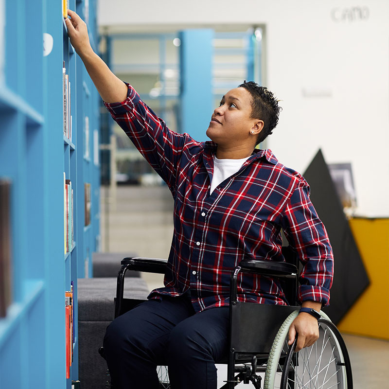 A woman sitting in a wheelchair reaching for a book on the shelf in a library
