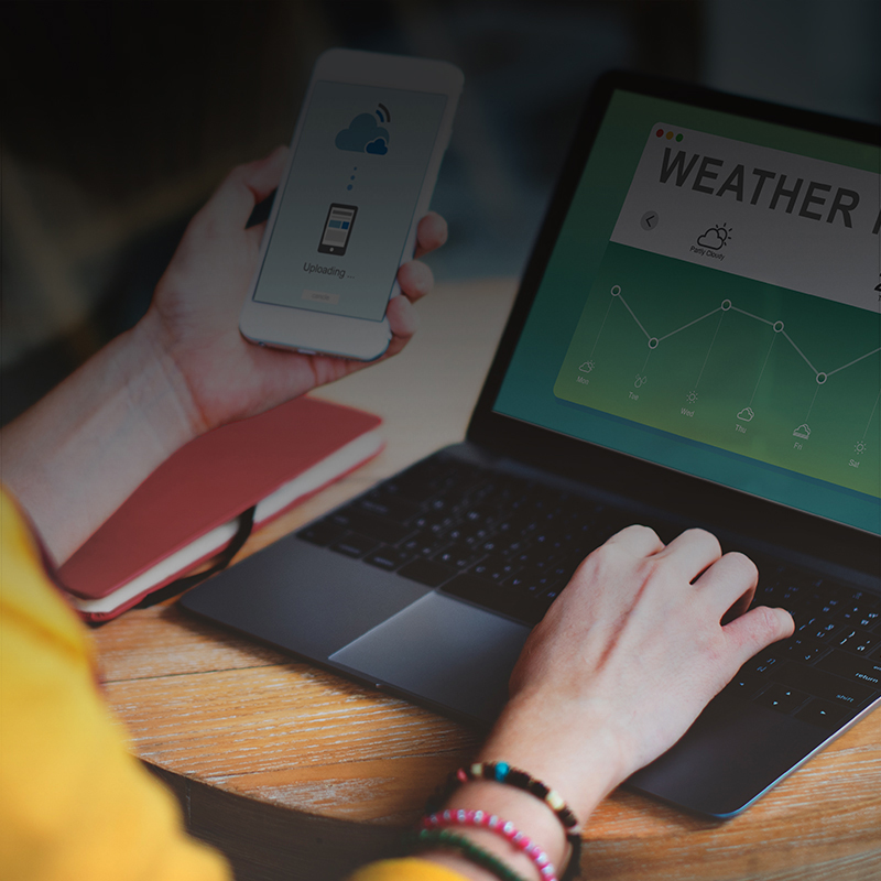 a photo of a woman wearing a yellow sweater looking at weather alerts on her computer and smart phone.