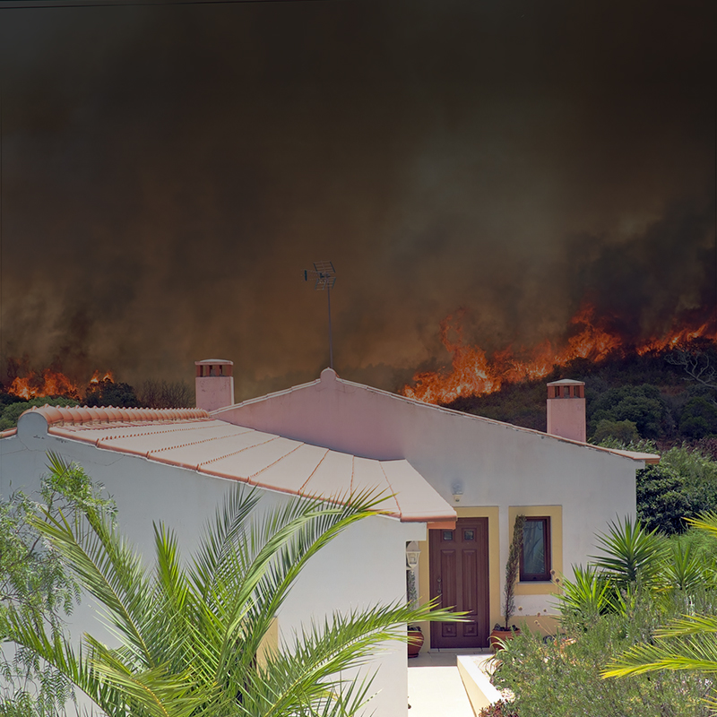 Image showing a wildfire raging in the background. In the foreground is a white houes with a Spanish style roof.