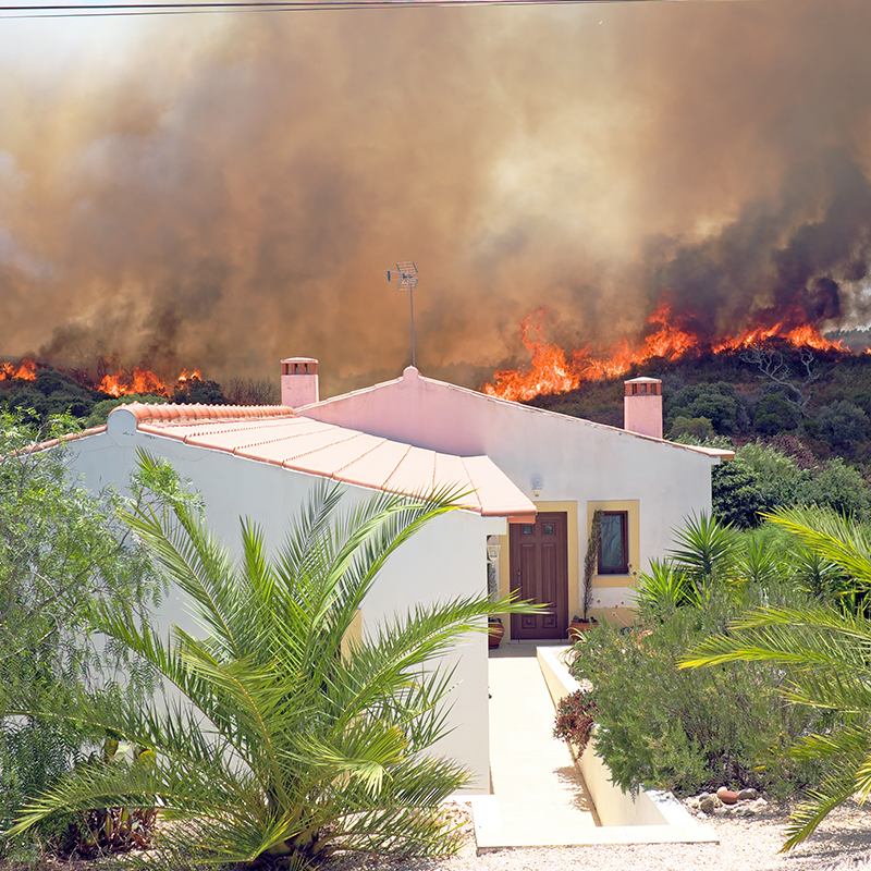 Image showing a wildfire raging in the background. In the foreground is a white houes with a Spanish style roof.